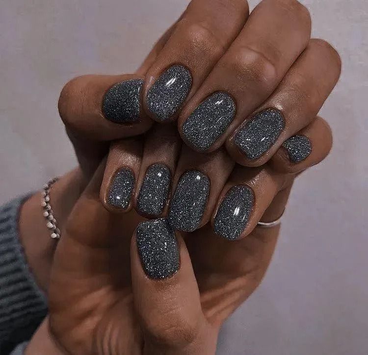 Hands with short, oval-shaped nails painted with sparkling, dark gray glitter nail polish.