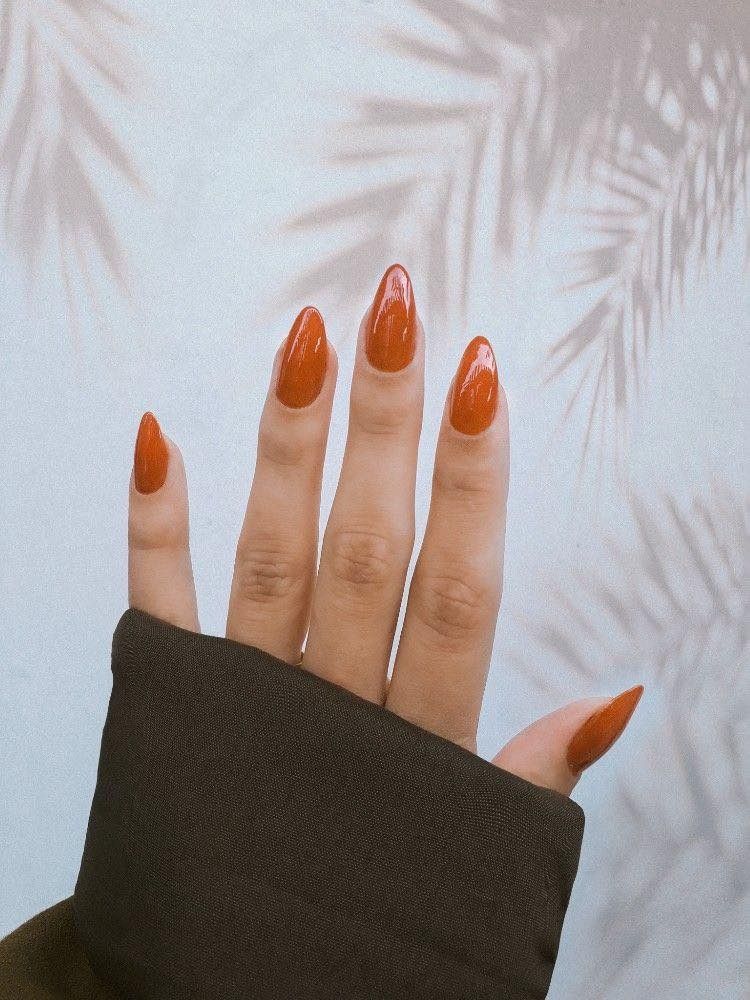 Hand with long, pointed, orange nails. Wearing a dark sleeve, palm up, with shadows of leaves on the wall.