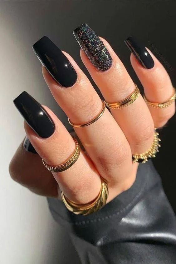 Hand with long, glossy black nails, one glitter nail, wearing multiple gold rings.