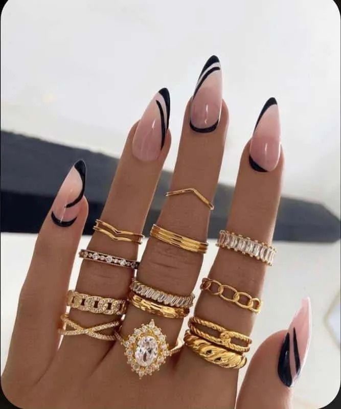 Hand with long, pointed nails in nude and black nail art. Multiple gold rings adorn fingers.