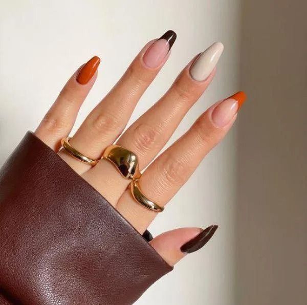 Hand with gold rings and long, oval-shaped nails painted in shades of brown, beige, and orange.