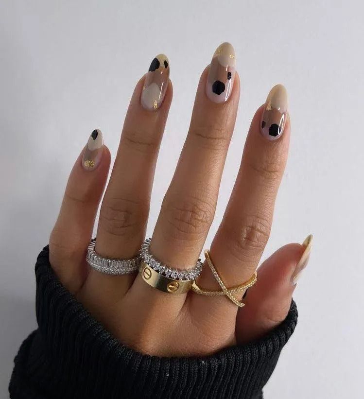 Hand with almond-shaped nails in beige tones, adorned with black spots, wearing multiple rings on a black sweater.
