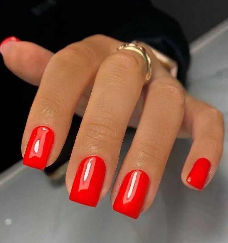 Hand with bright red square-shaped polished nails, wearing a gold ring.