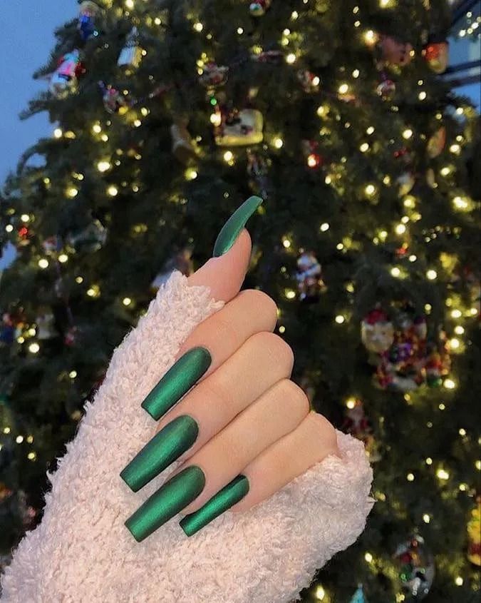 Hand with long, matte green nails in front of a decorated Christmas tree with lights.