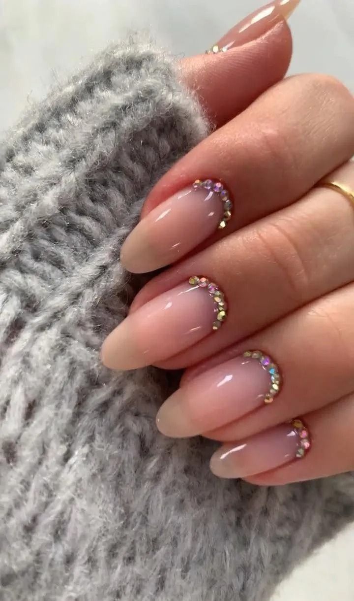 Close-up of a hand with oval-shaped nails, french tips, and rhinestones. The hand is resting on a gray knit fabric.