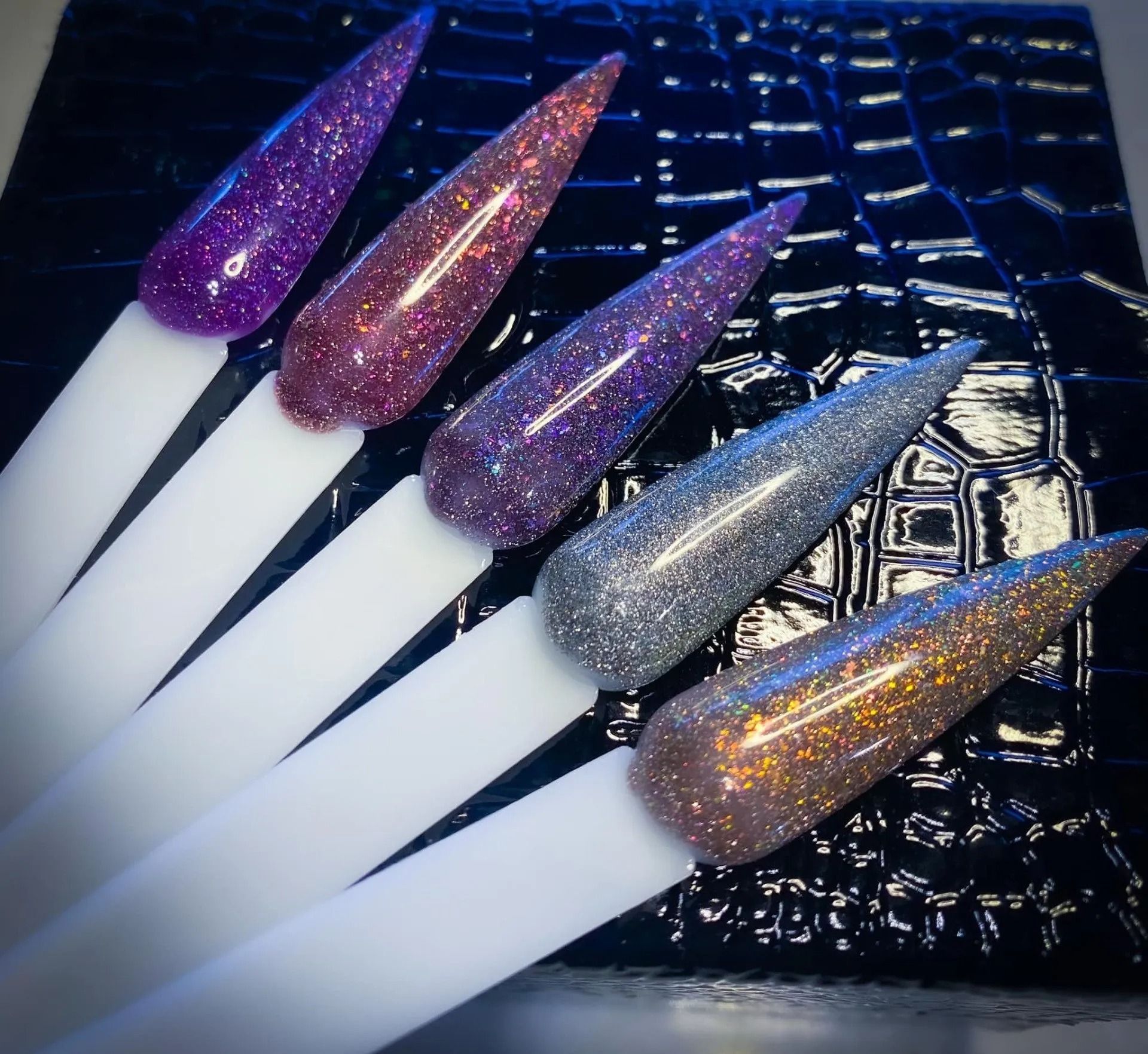 Five glitter nail polish swatches on white sticks, in purple, rose, lavender, silver, and gold against a textured black background.