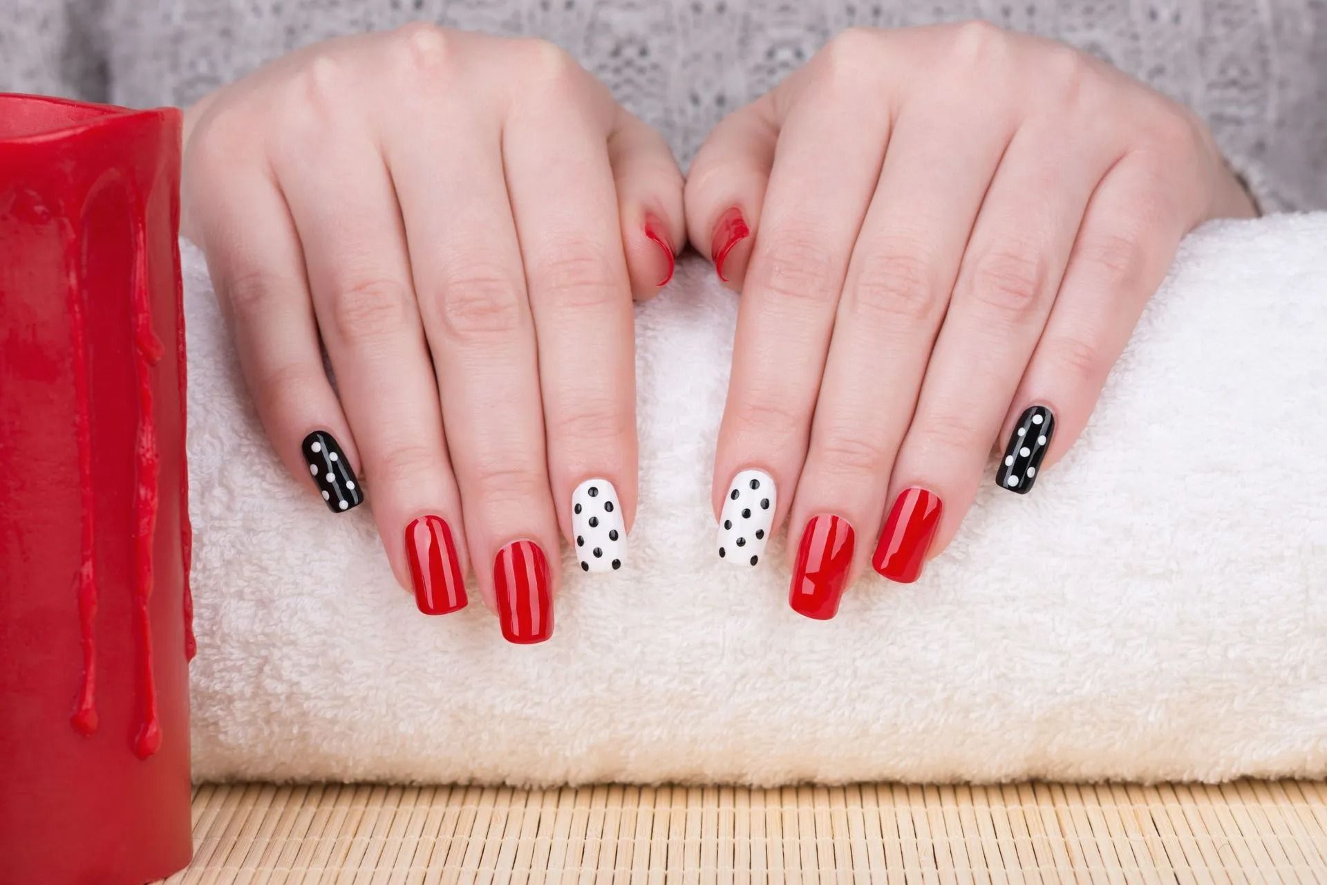 Hands with red, black and white polka-dot nail art resting on a white towel, next to a red candle.