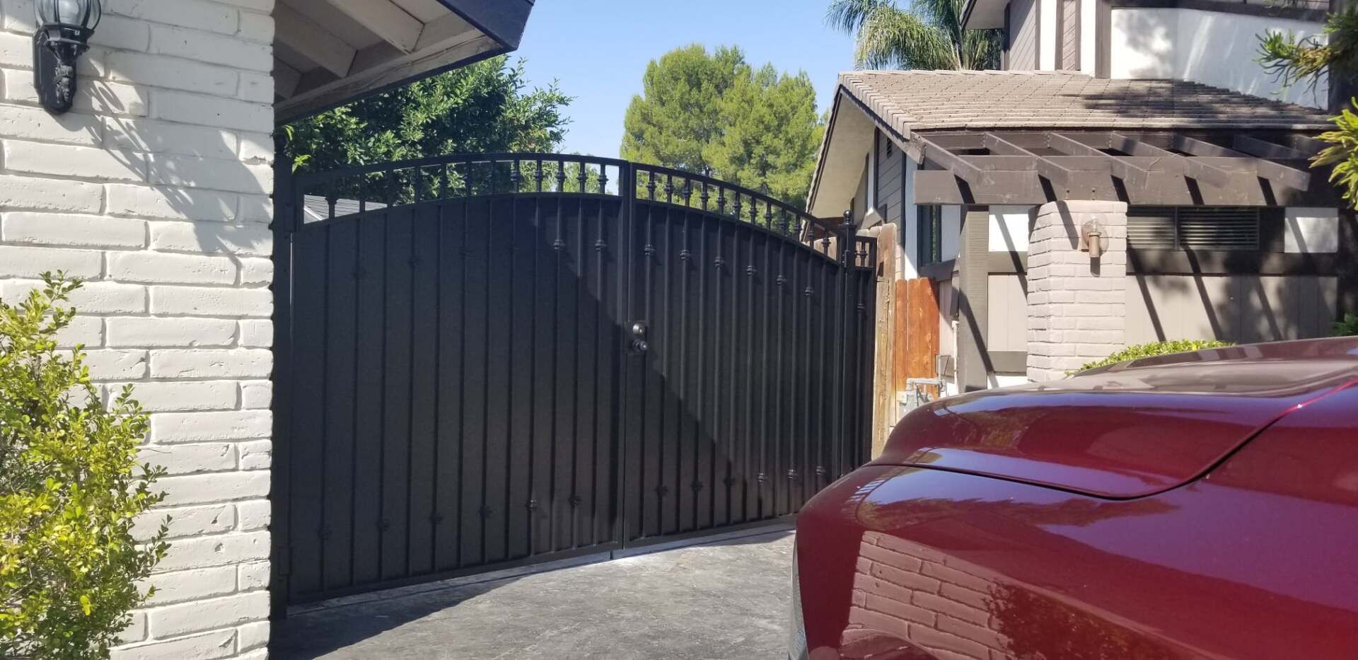 Black gate with driveway, brick house, red car, trees, and blue sky.
