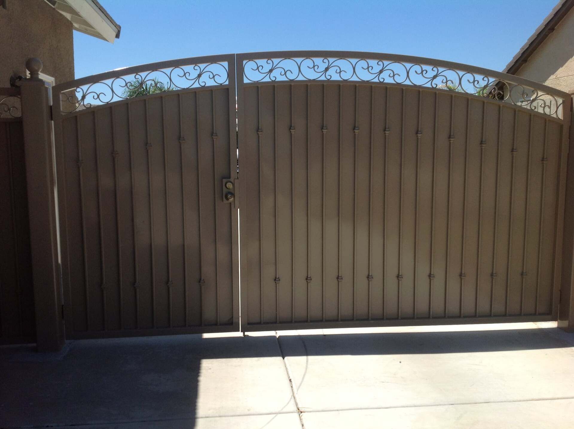 Tan wooden gate with wrought iron detailing, set in concrete, under a clear blue sky.