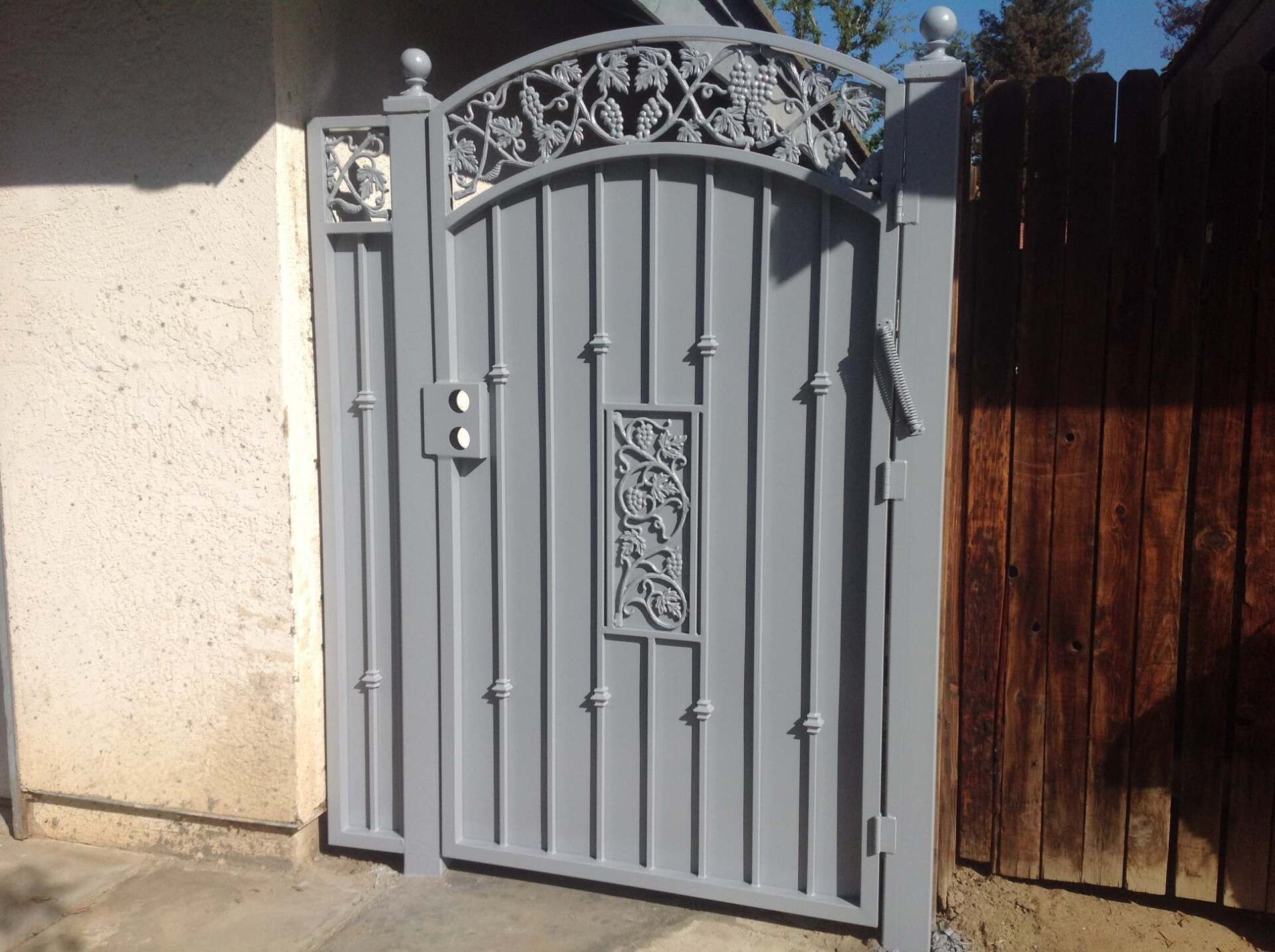 Gray metal gate with decorative vine detailing, set against a concrete wall and wooden fence.