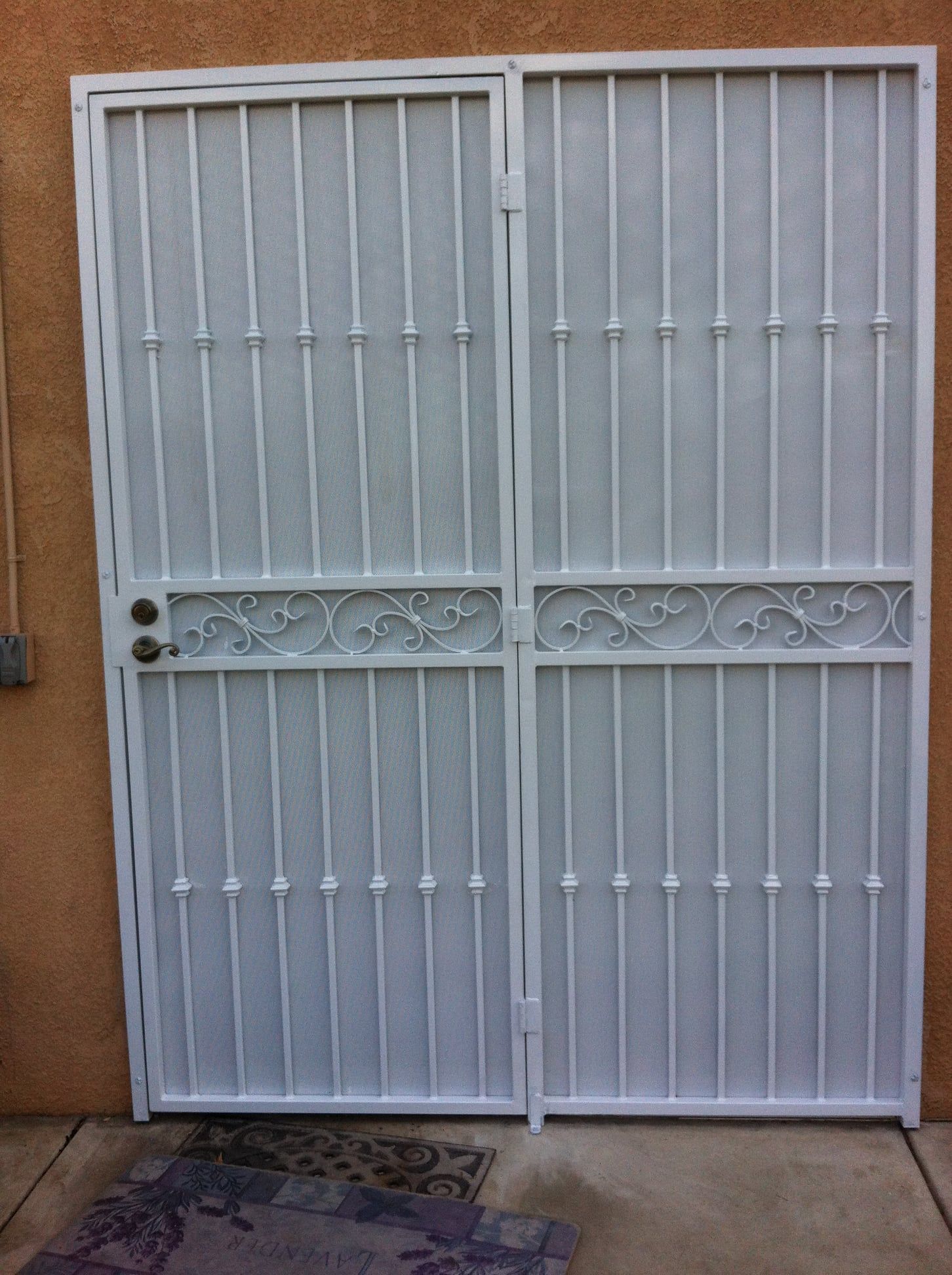 White metal security doors with decorative scrollwork against an orange wall and concrete floor.