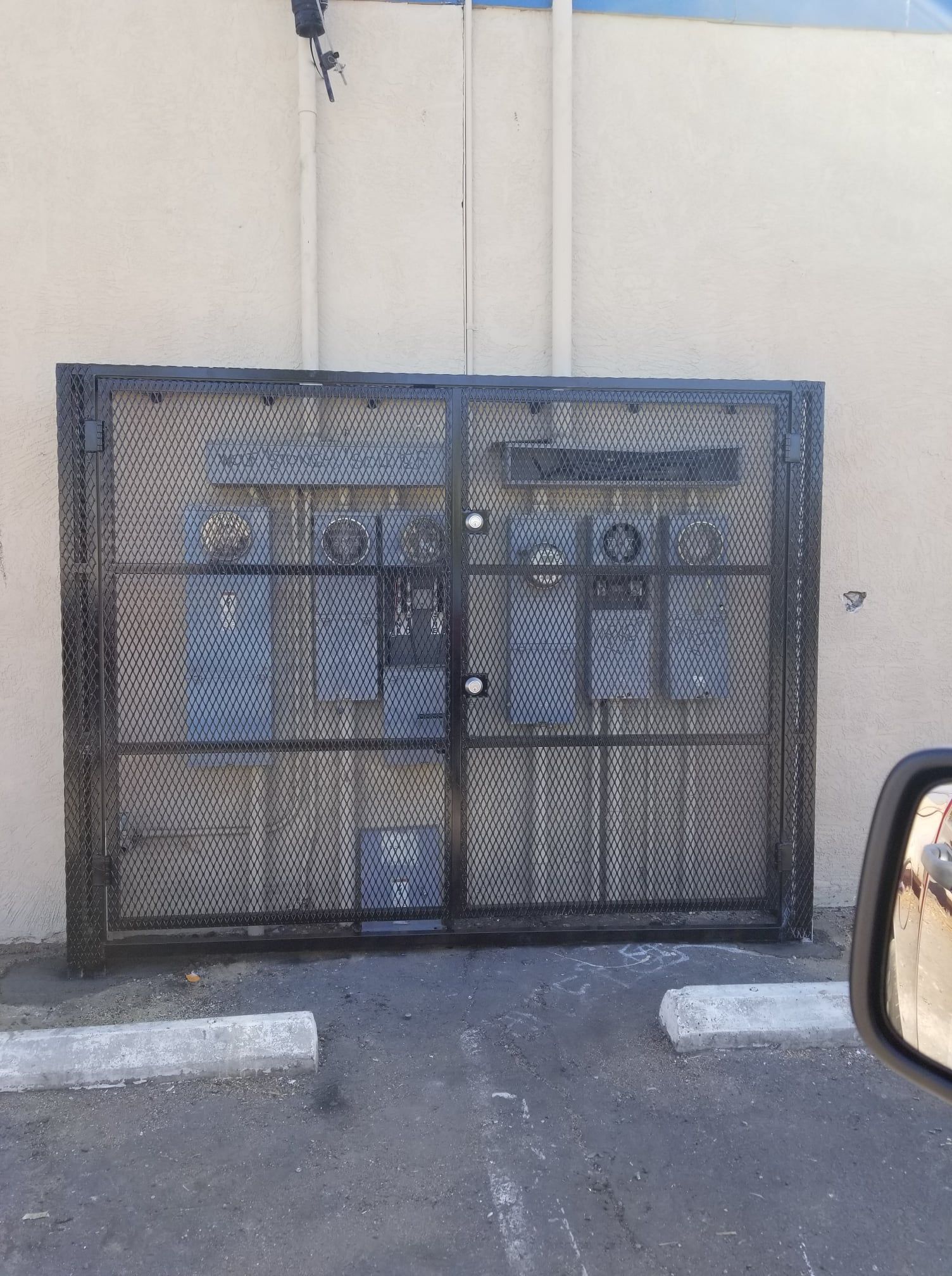 Black metal cage enclosing electrical equipment against a beige wall, parking area in front.