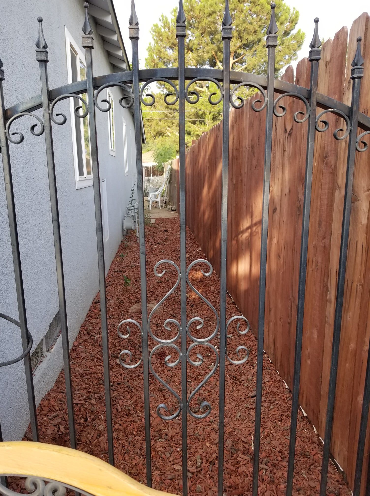 Black wrought iron gate open, leading to a narrow pathway with red mulch, bordered by a wooden fence and gray building.