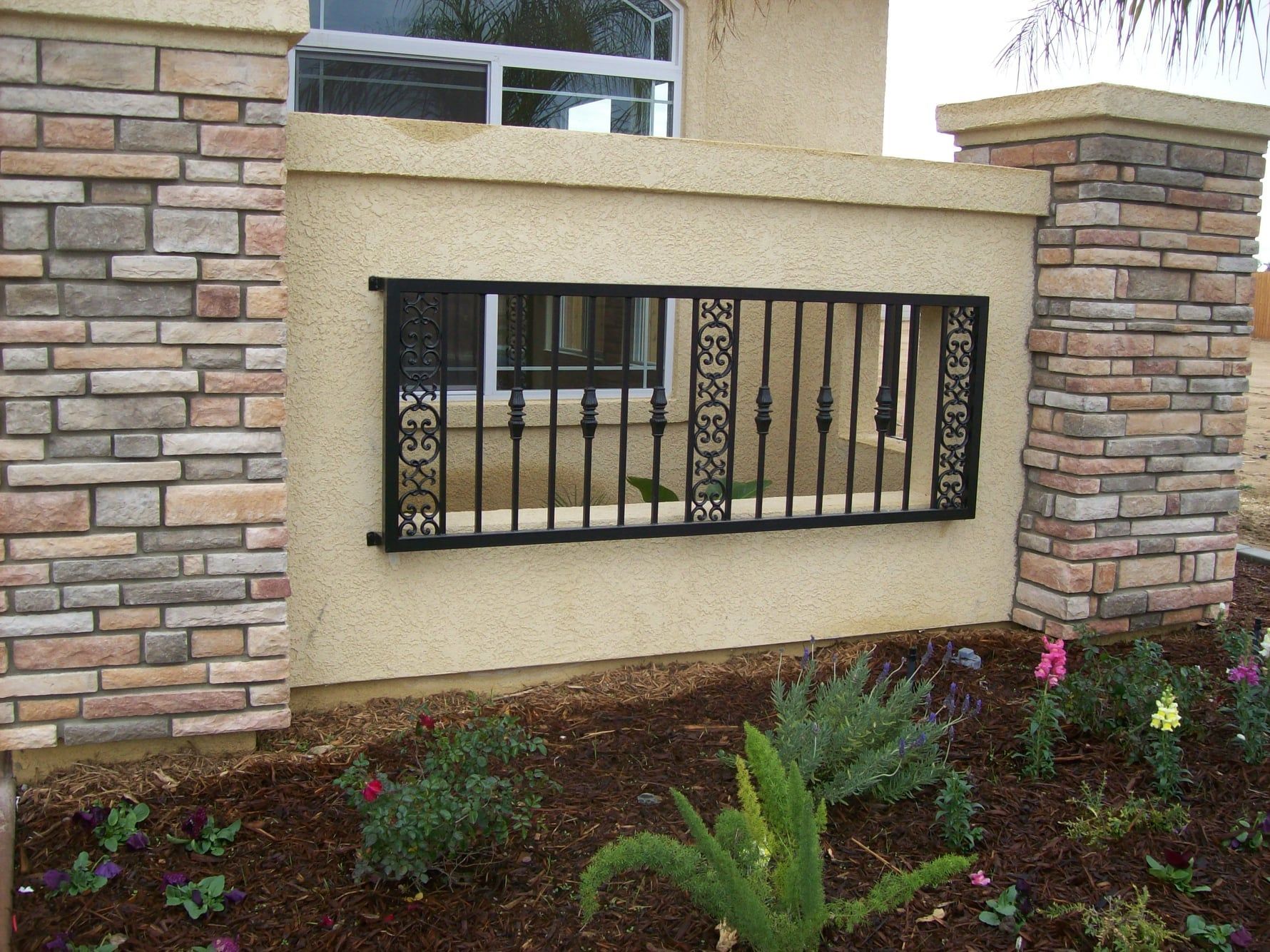 Decorative wrought iron window security bars on a stucco wall, flanked by stone columns, with a flower bed in front.
