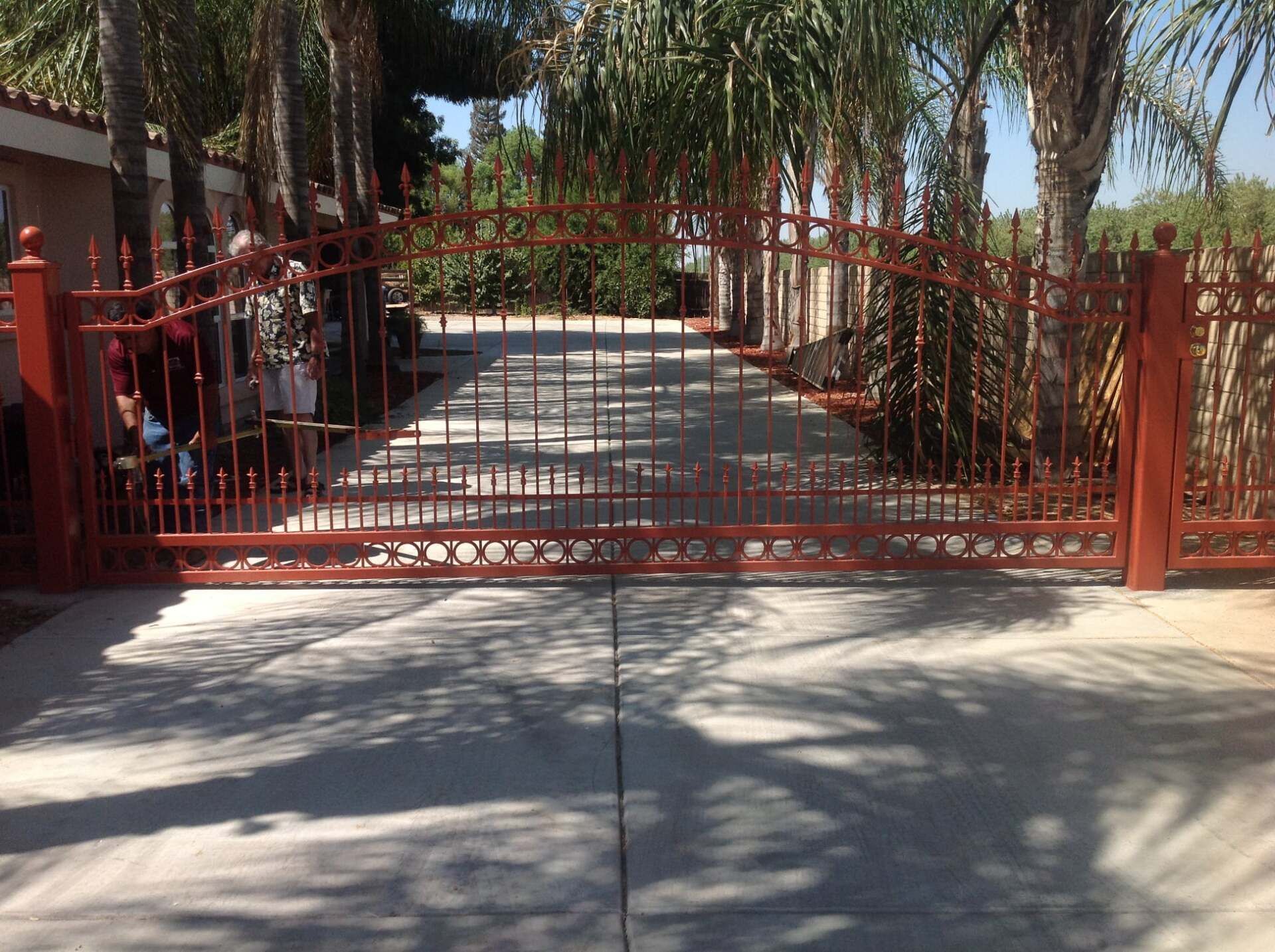 Red metal driveway gate, open, with concrete driveway and palm trees in background.