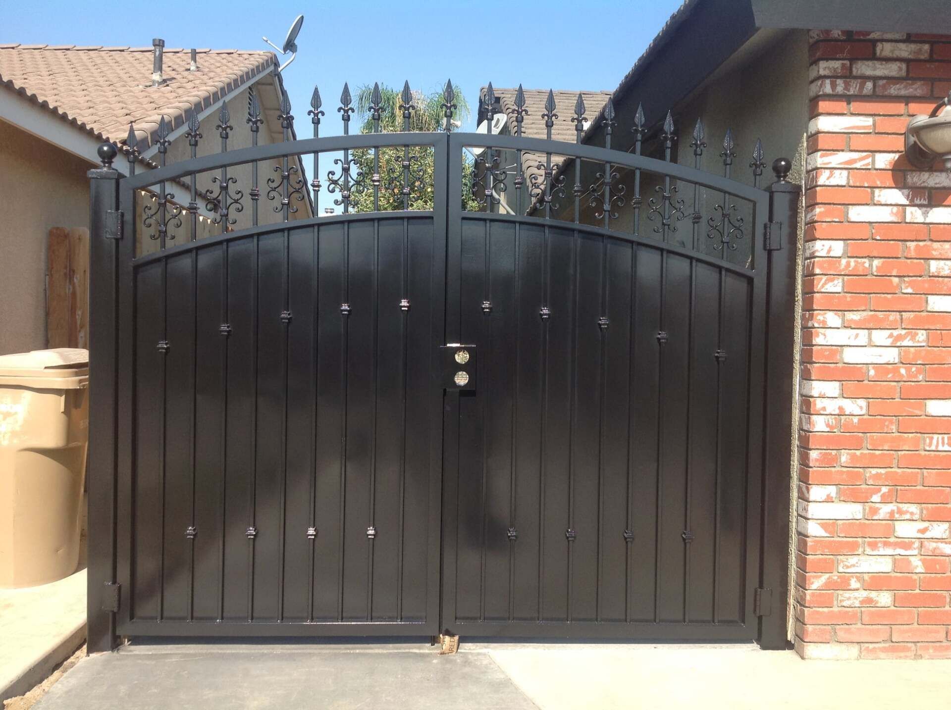 Black metal driveway gate with vertical wooden slats and decorative top, against a brick and stucco house.