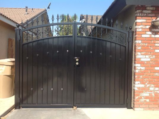 Black metal driveway gate with vertical wooden panels and decorative top, set between two structures.