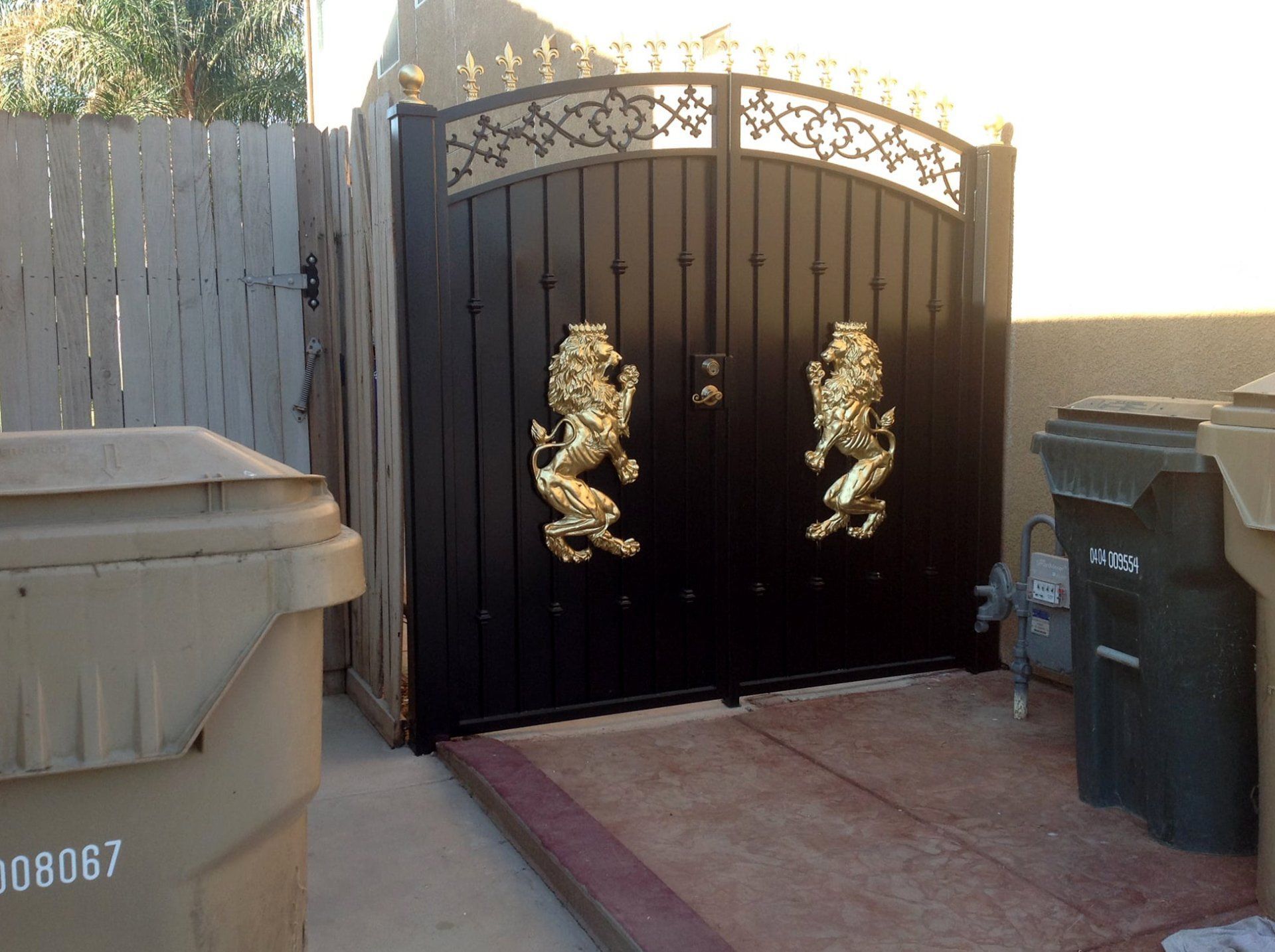 Black gate with gold lion door knockers, two trash cans on either side.