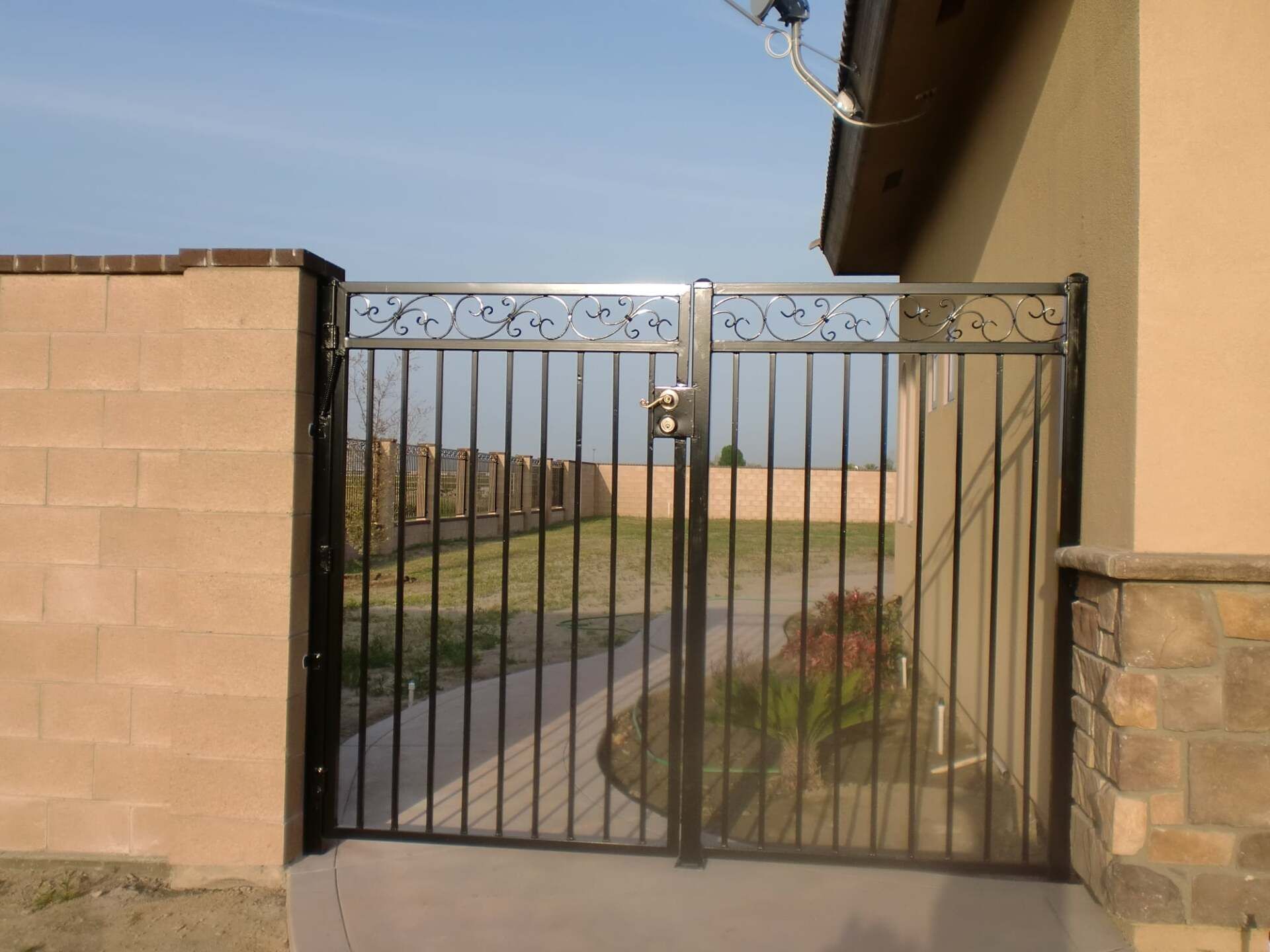 Black metal gate in a yard, between a brick wall and a stucco house with a satellite dish.