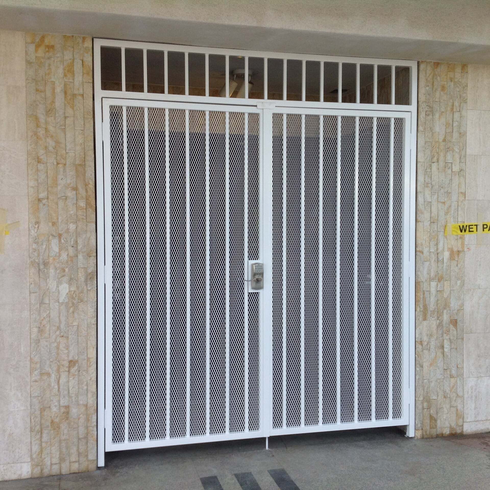 White metal security gate with vertical bars and intricate design, set between textured beige walls.