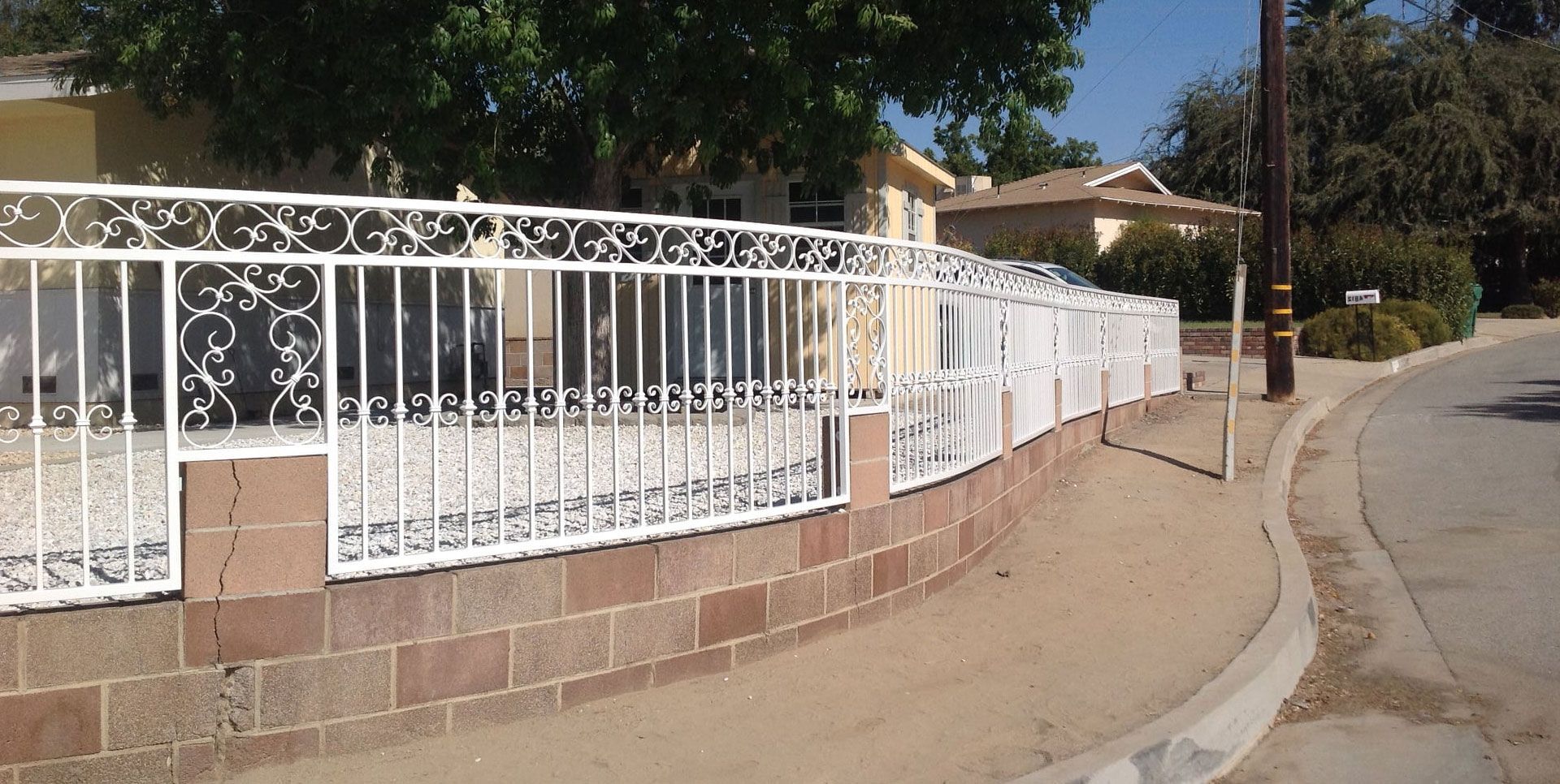 White ornate fence on a brick wall curves along a sandy sidewalk next to a street.