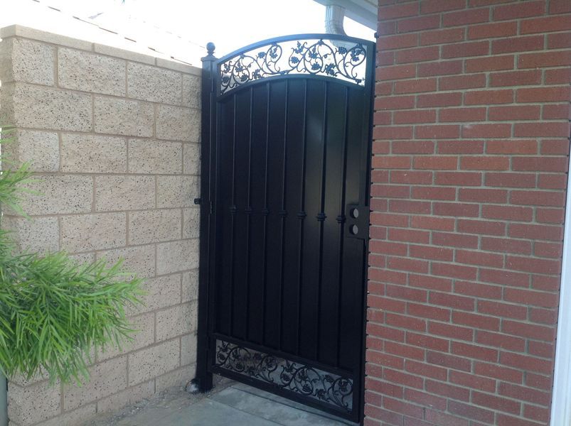 Black metal gate set between a tan brick wall and a red brick wall, with decorative scrollwork.
