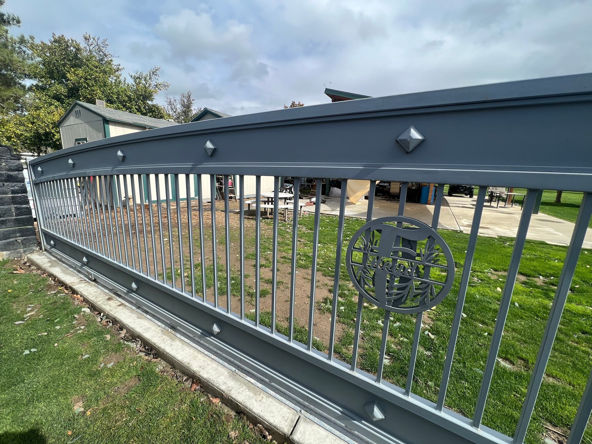 Gray metal fence with decorative diamond accents, vertical bars, and circular emblem, in front of greenery and a building.