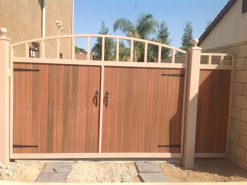 Wooden double gate with arched metal top, brown wood, tan frame, against a sunny backdrop.