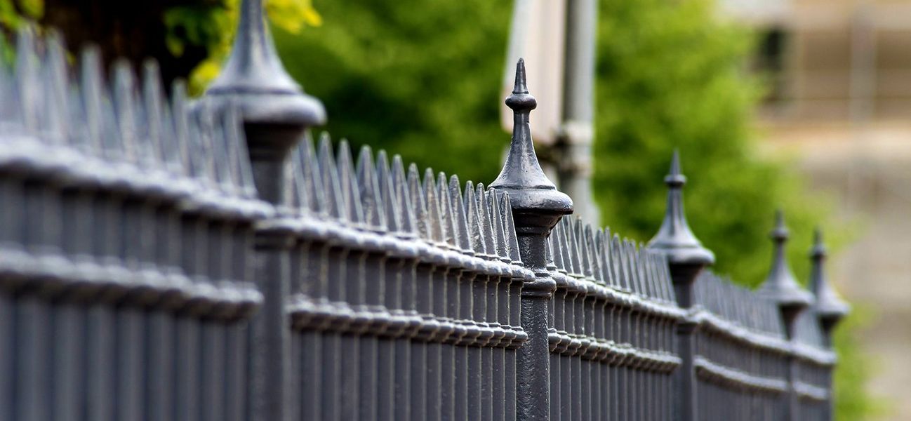 Black wrought iron fence with decorative finials, green foliage in background.