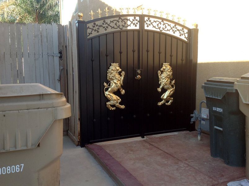 Black gate with gold lion door knockers, decorative top, flanked by trash bins and a wooden fence.