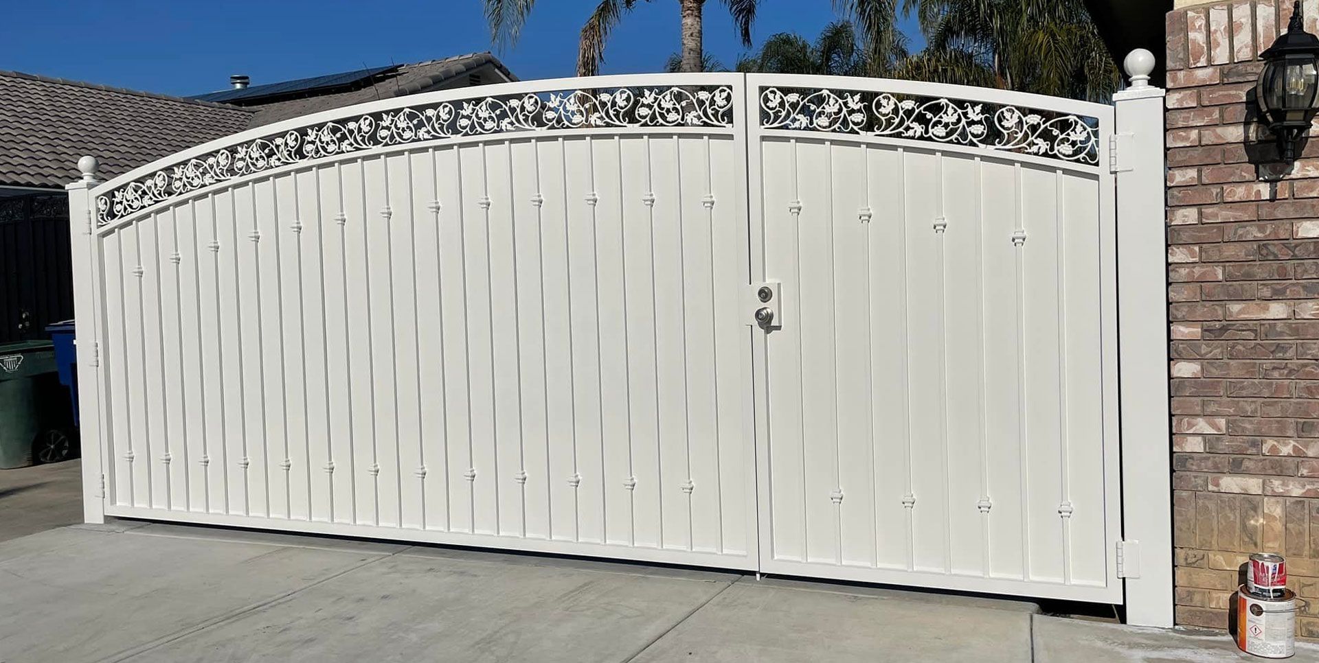 White arched gate with decorative scrollwork, set in front of a brick pillar.