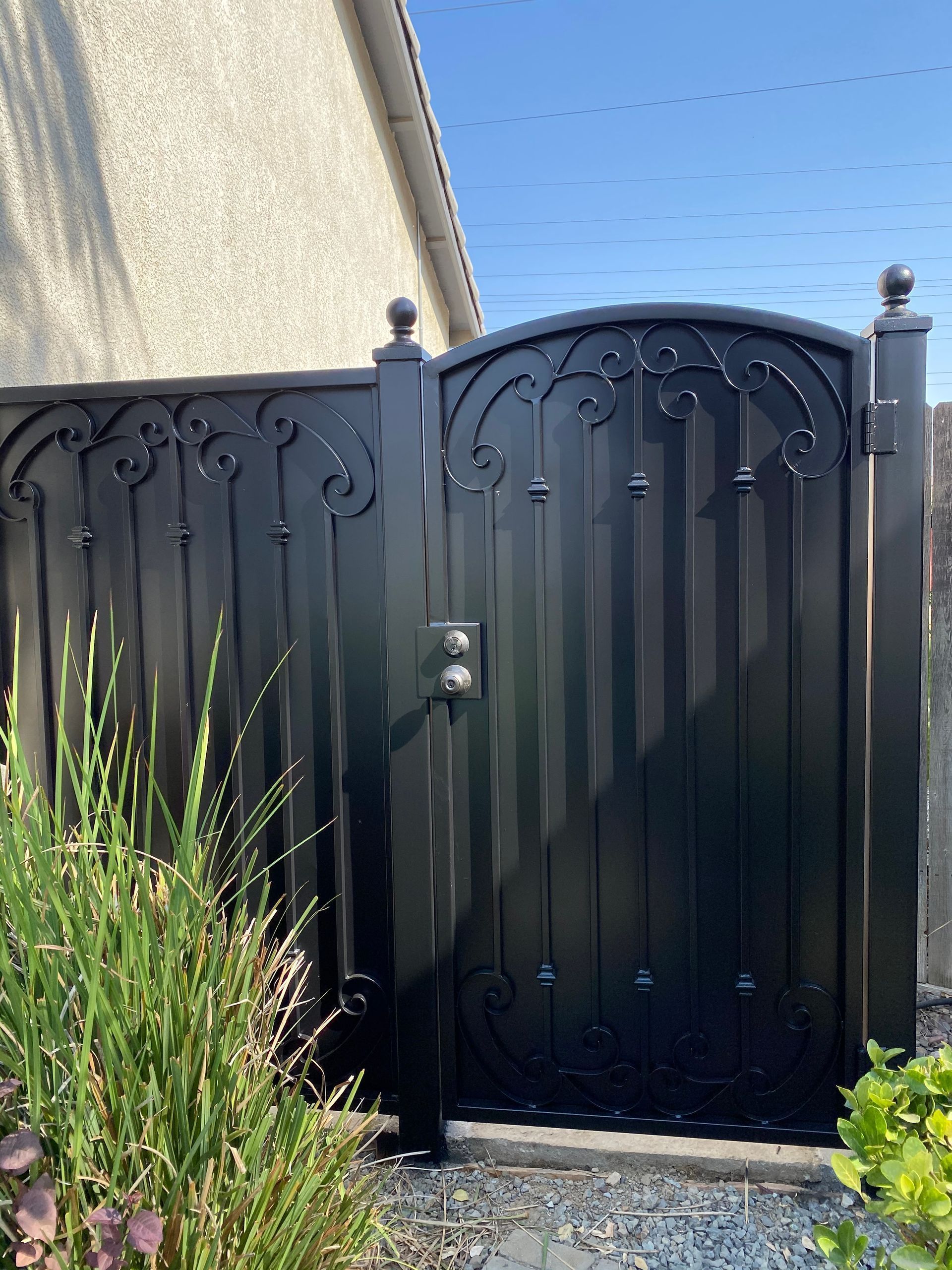 Black metal gate with ornate details, between two pillars, in front of a light-colored wall.