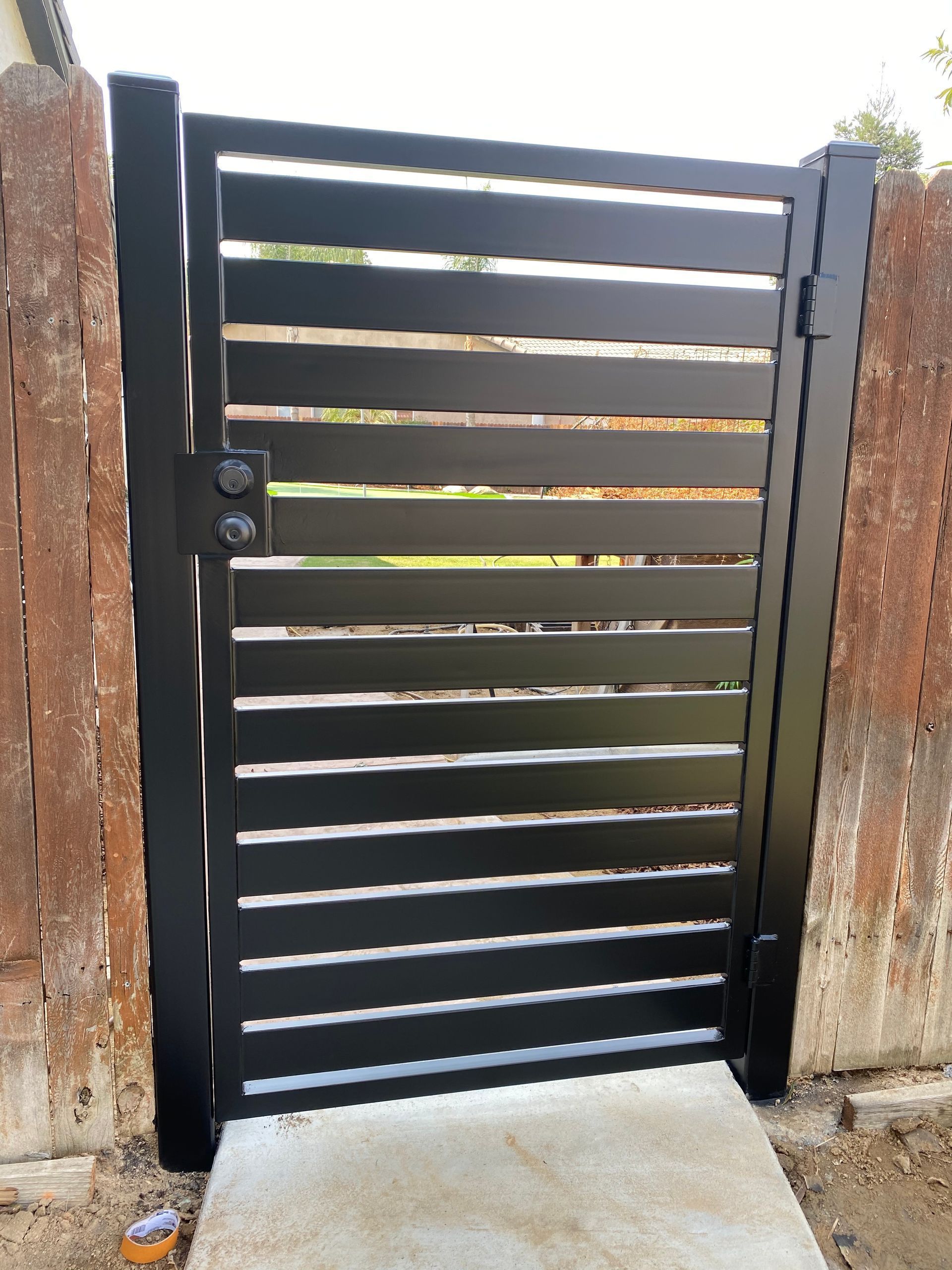 Black horizontal slat gate set in a concrete path, between wooden fence posts.