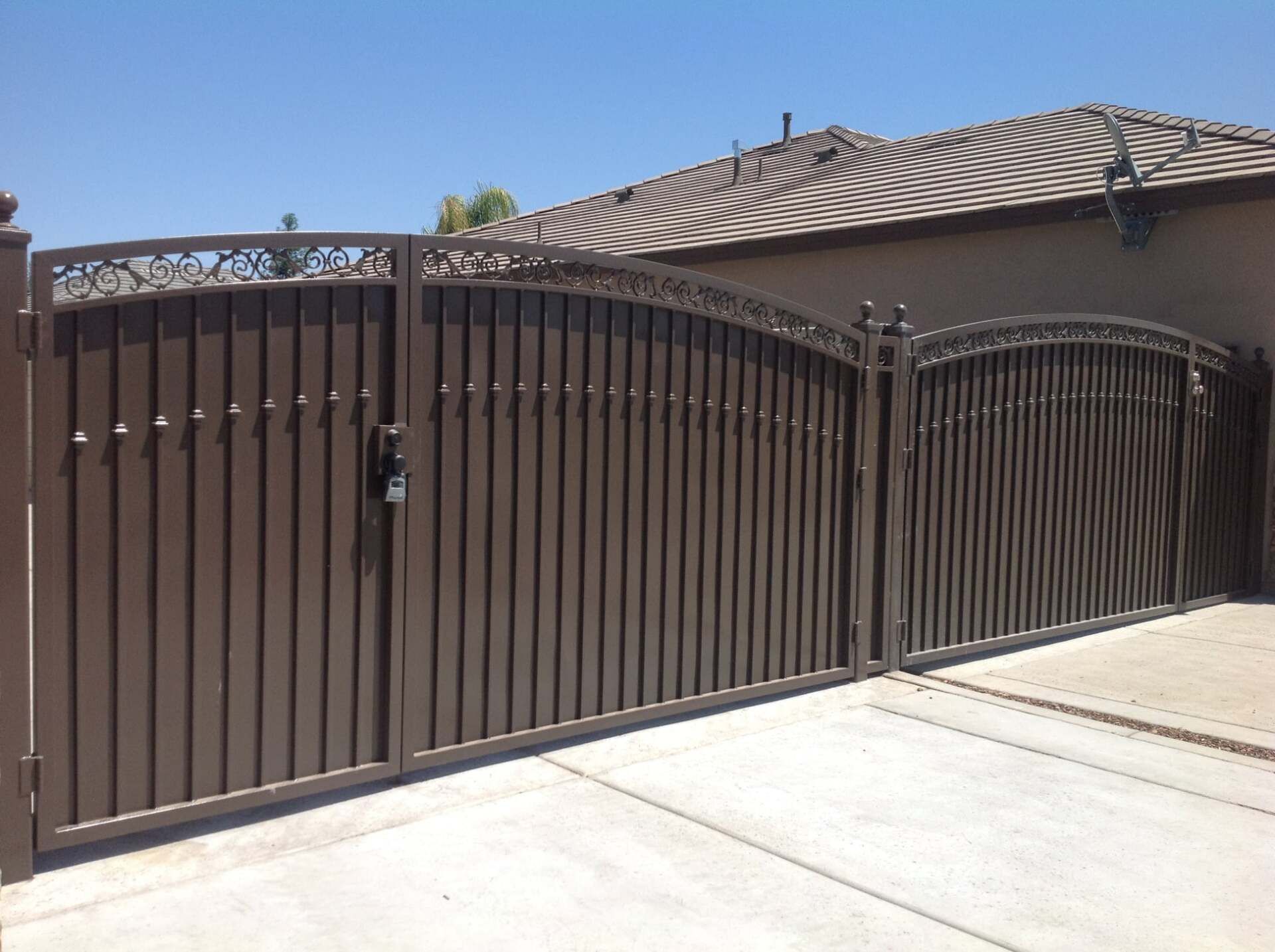 Brown metal driveway gate with ornate top, in front of a tan building, on a sunny day.