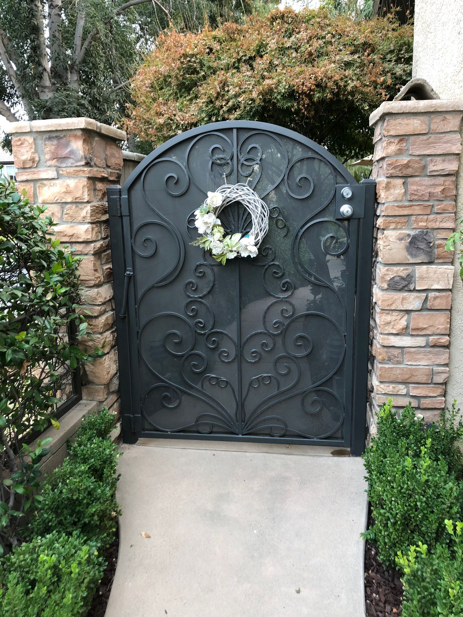 Ornate black wrought iron gate with a white floral wreath, flanked by brick columns and hedges.