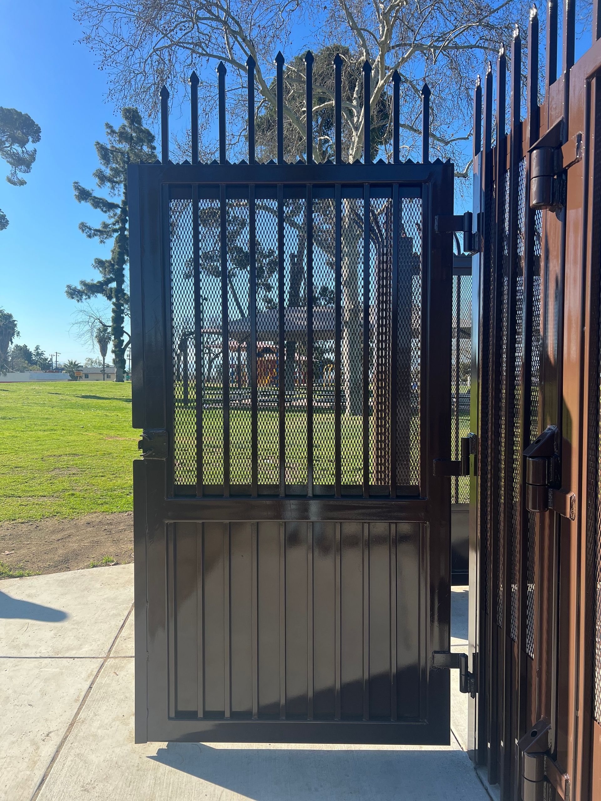 Black metal gate, partly open, reveals a grassy park on a sunny day.