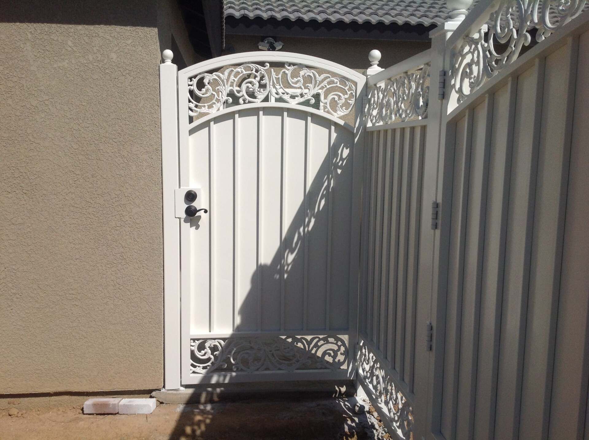 White metal gate with ornate scrollwork, attached to a matching fence, next to a stucco wall.