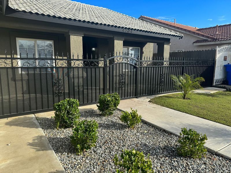 A house with a black metal fence, small bushes, and a pathway on a sunny day.