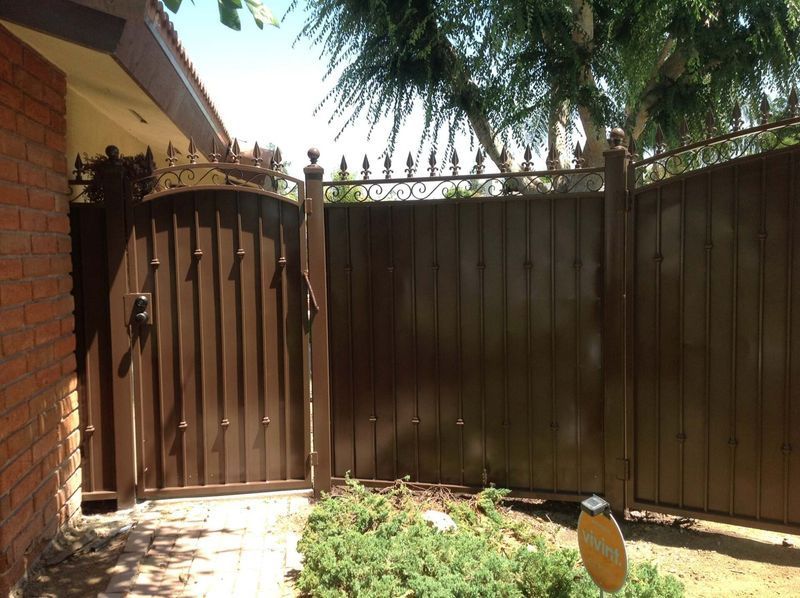 Brown metal gate and fence in front of a brick building and shrubbery, with a decorative wrought iron top.
