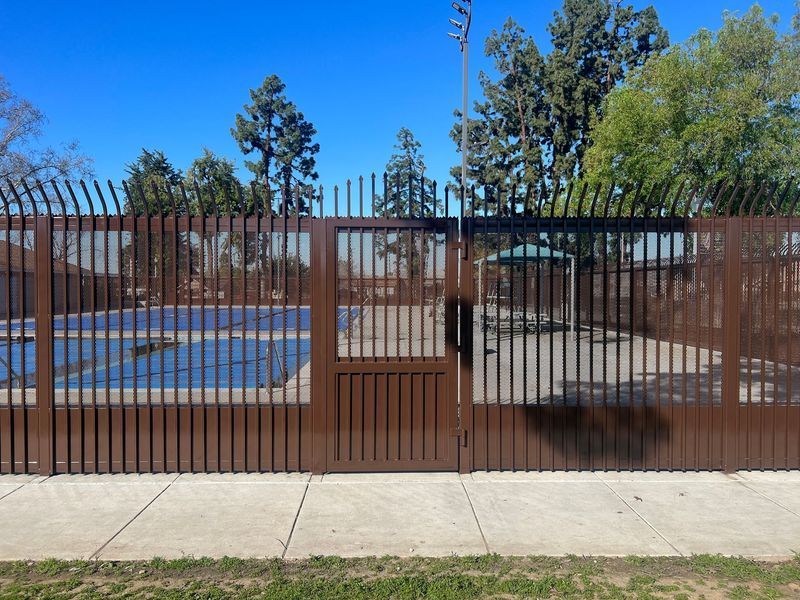 Brown metal fence with gate in front of a blue-covered pool, sidewalk and trees under a clear sky.