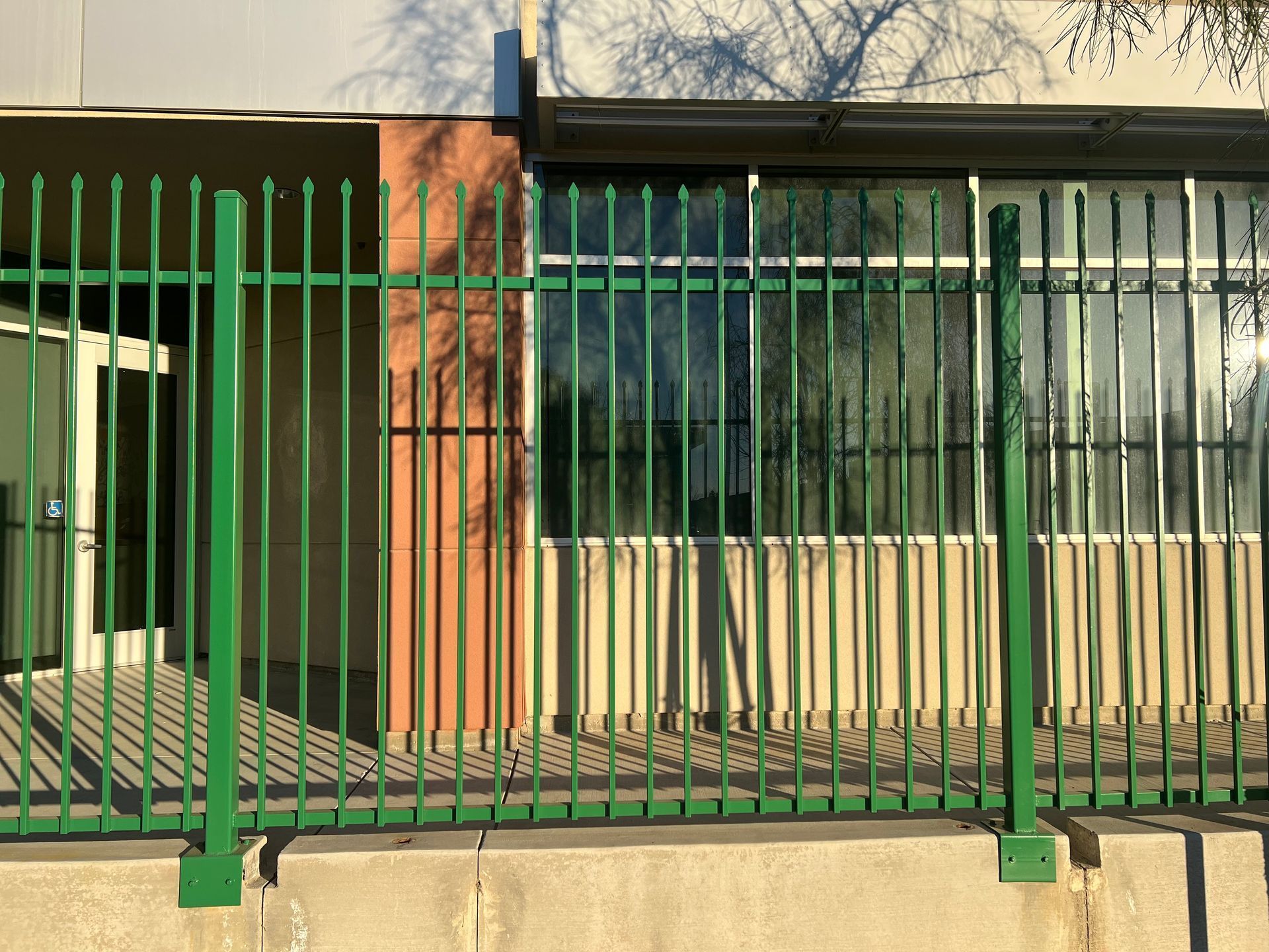 Green metal fence in front of a building with windows, casting long shadows.