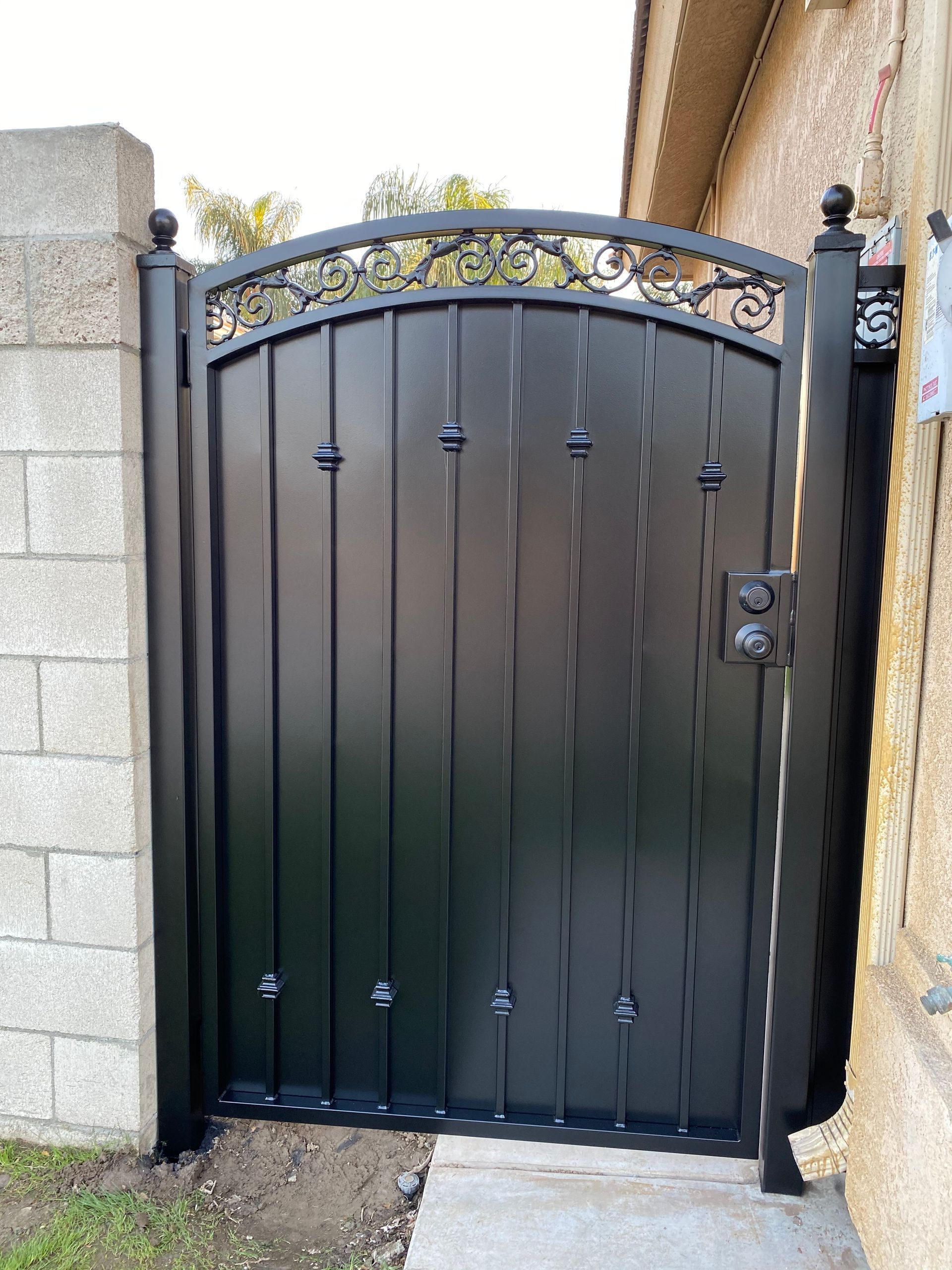 Black metal gate with arched top, ornate design, between concrete walls.
