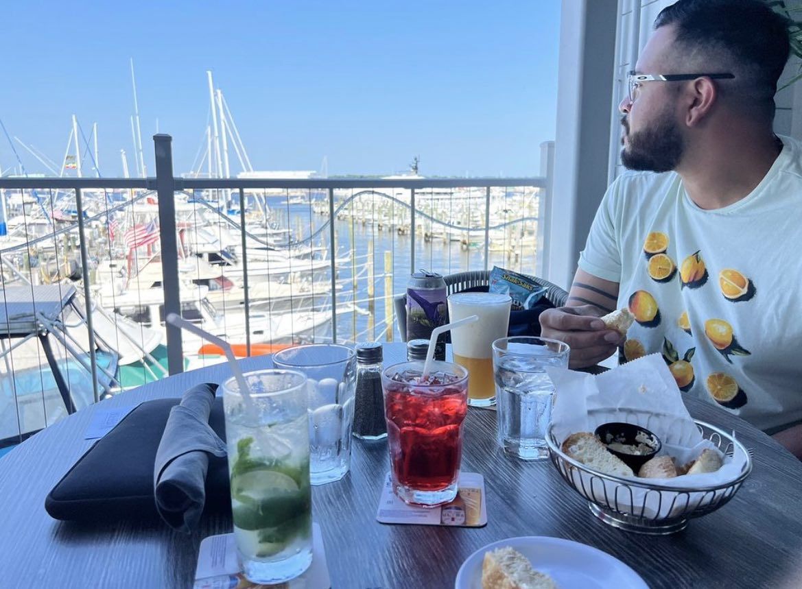 Man at outdoor table with drinks and bread, overlooking boats.