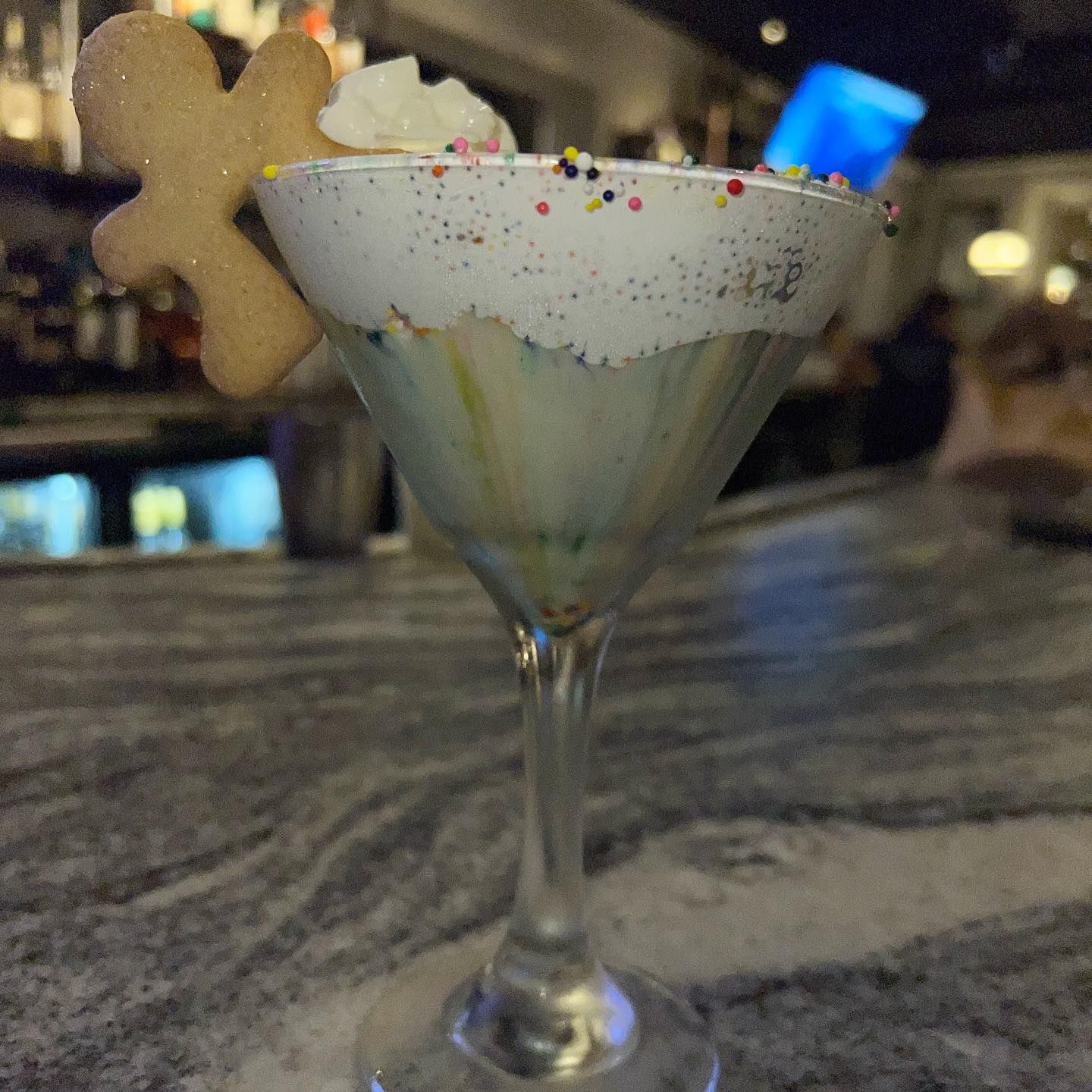 Martini glass with a dessert cocktail, gingerbread man cookie, sprinkles, and whipped cream on a marble bar.