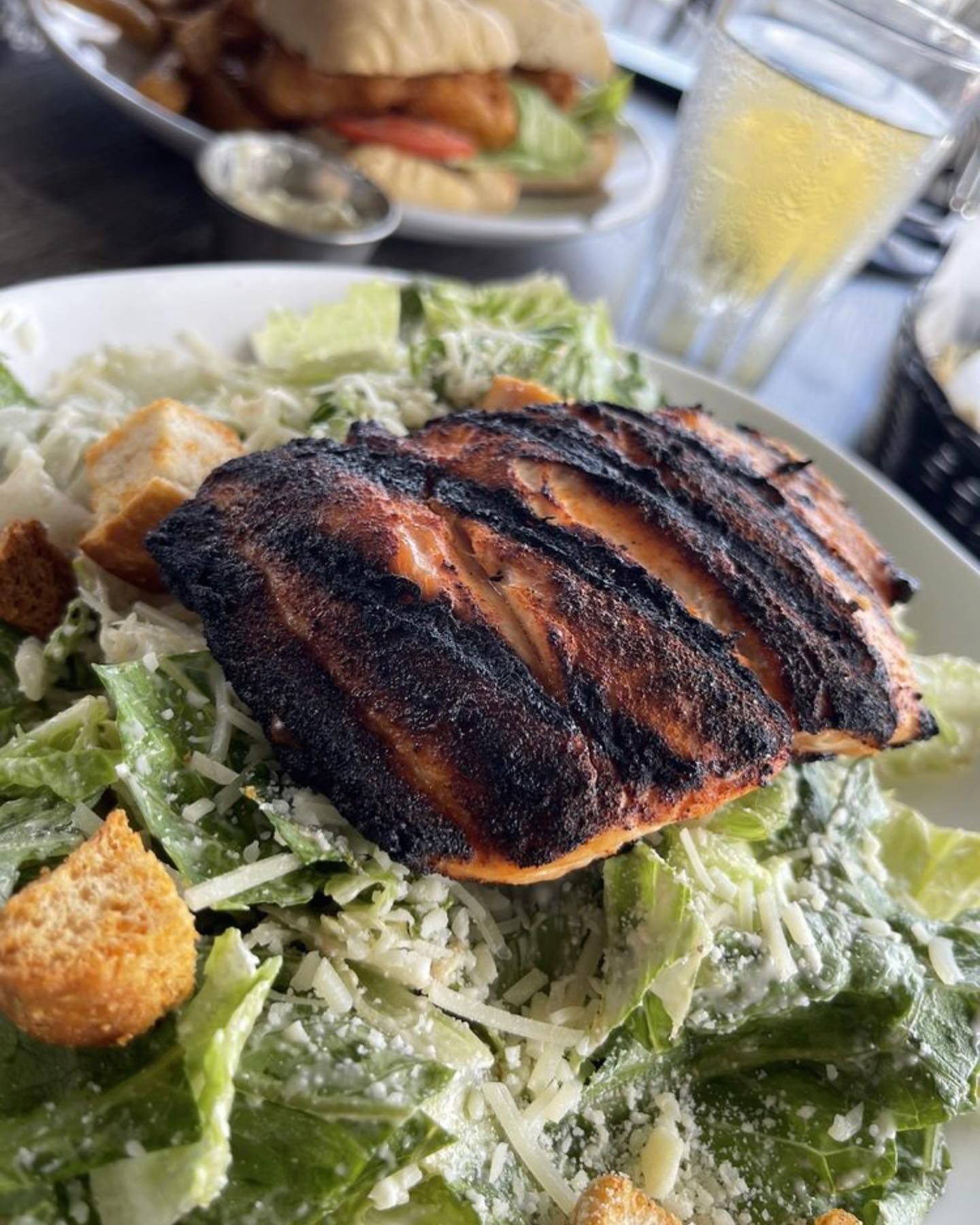 Grilled salmon atop Caesar salad with croutons. Fish sandwich and drink in the background.