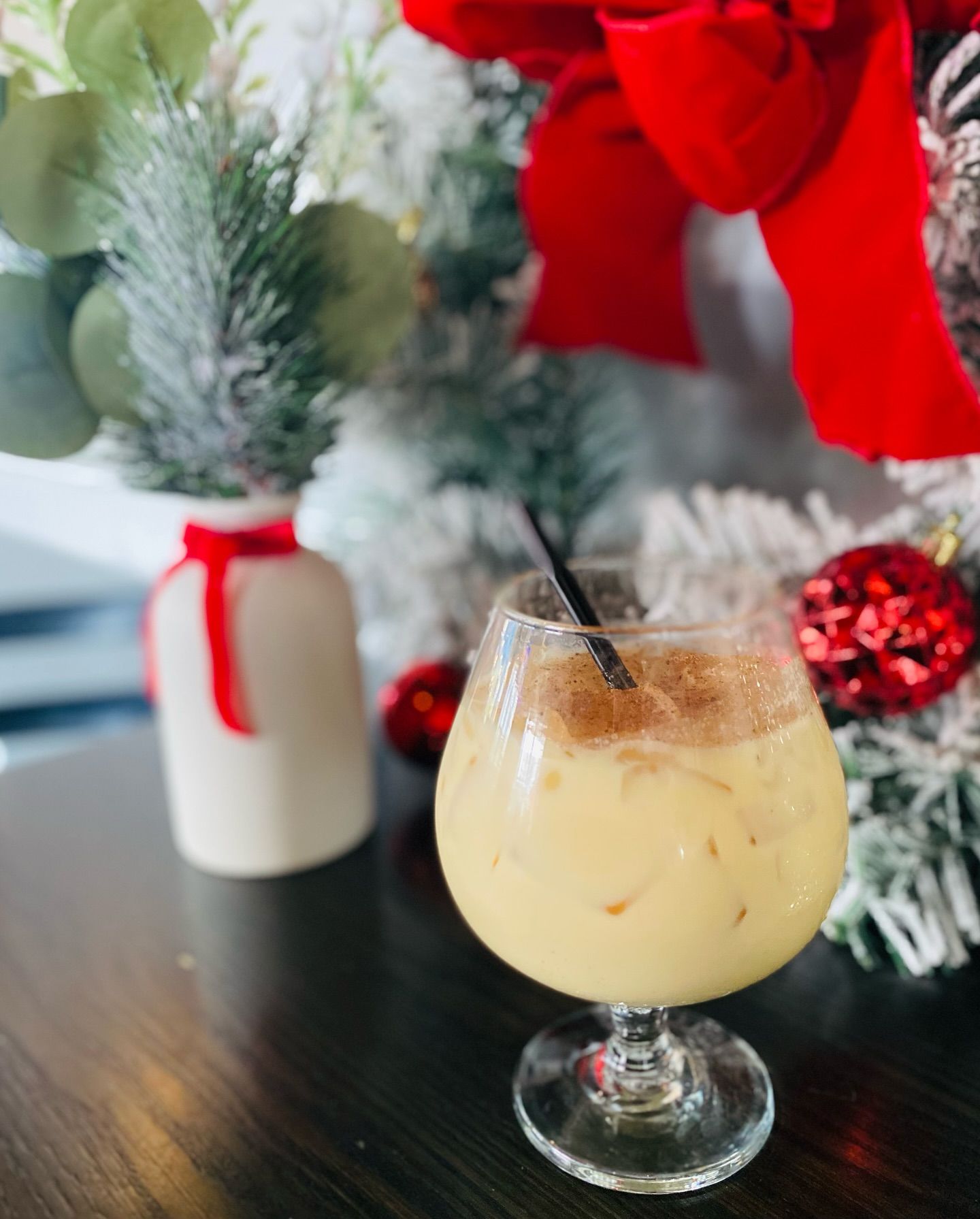 Eggnog cocktail in a glass with a straw, dusted with cinnamon, on a table, with Christmas decorations in the background.
