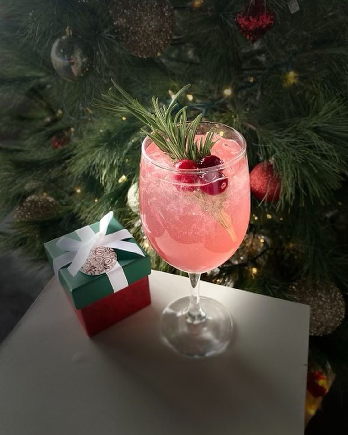 Cocktail in a glass with cranberries and rosemary, next to a wrapped gift, in front of a Christmas tree.