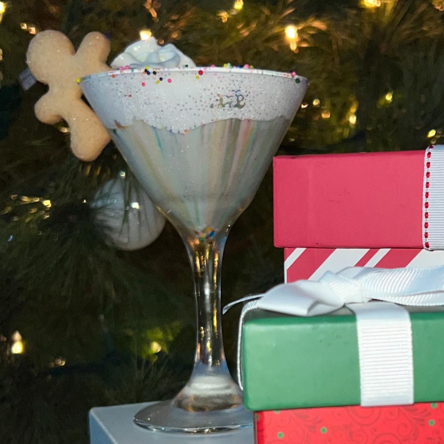 Martini glass with whipped cream and sprinkles, gingerbread cookie, next to Christmas presents and tree.