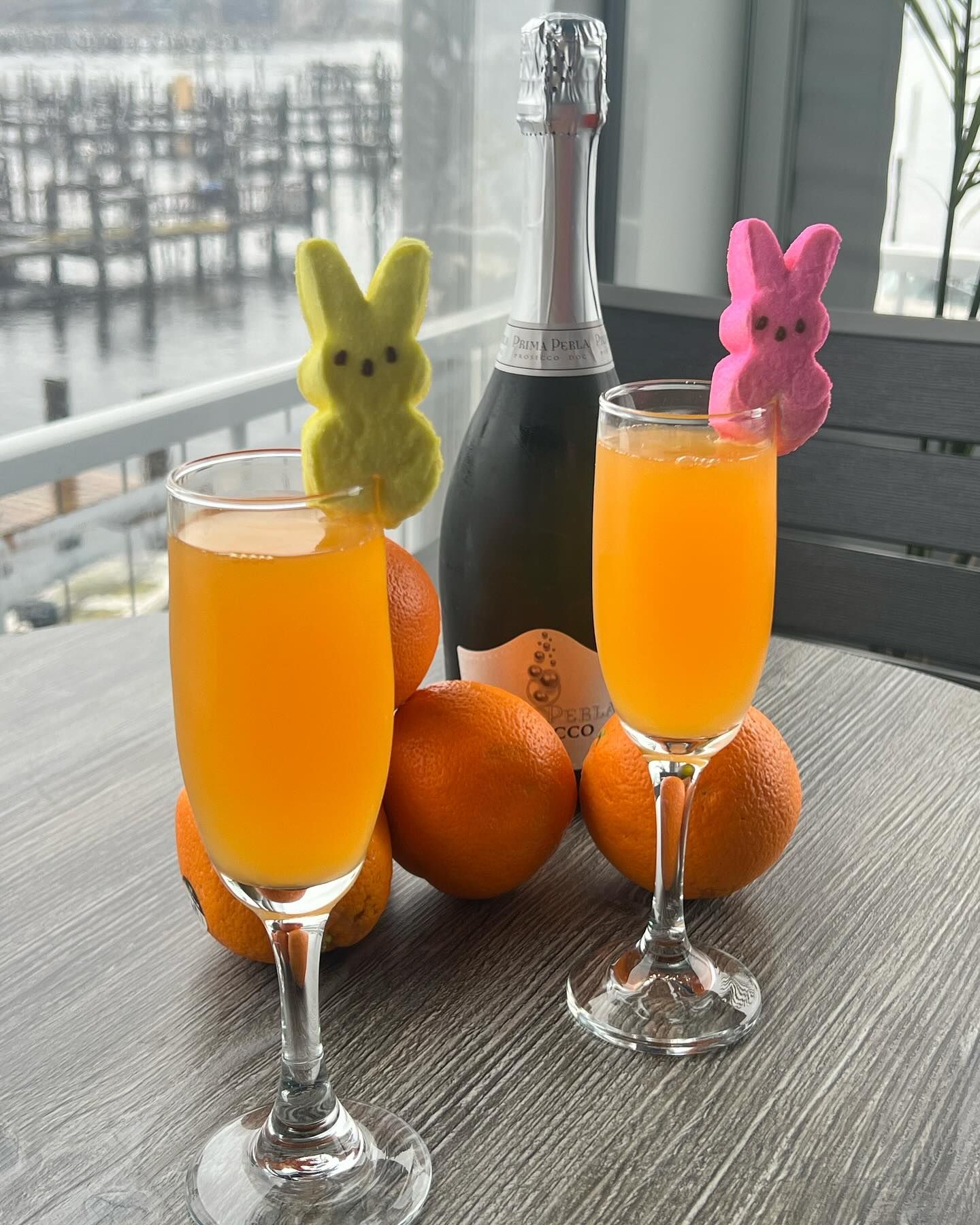 Two mimosa glasses with orange juice, champagne, oranges, and Peeps, on a table. Waterfront background.