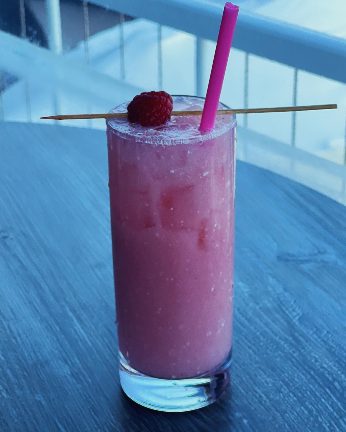 Pink cocktail in tall glass with raspberry garnish and pink straw.